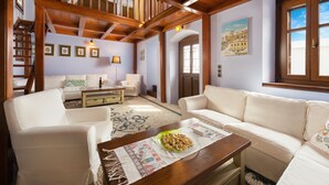A spacious living room with a high ceiling, wooden beams, a staircase, a white sofa, a coffee table with a decorative plate, and a door leading to another room. - Residenza Maria Lindos Villa (Rhodes)