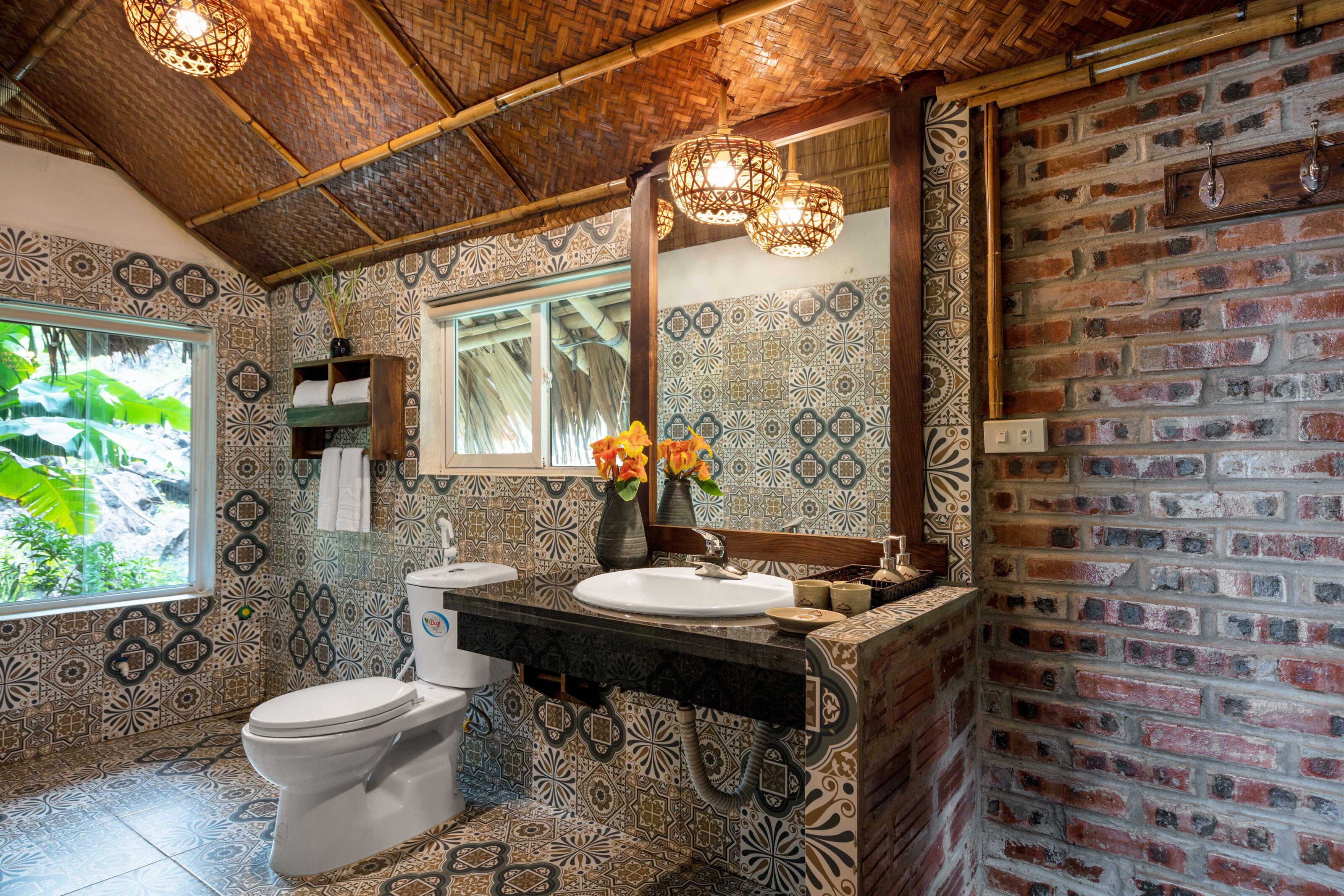 Un baño con pared de ladrillo, baldosas estampadas, un inodoro blanco, un lavabo con espejo y una ventana con vista a follaje.