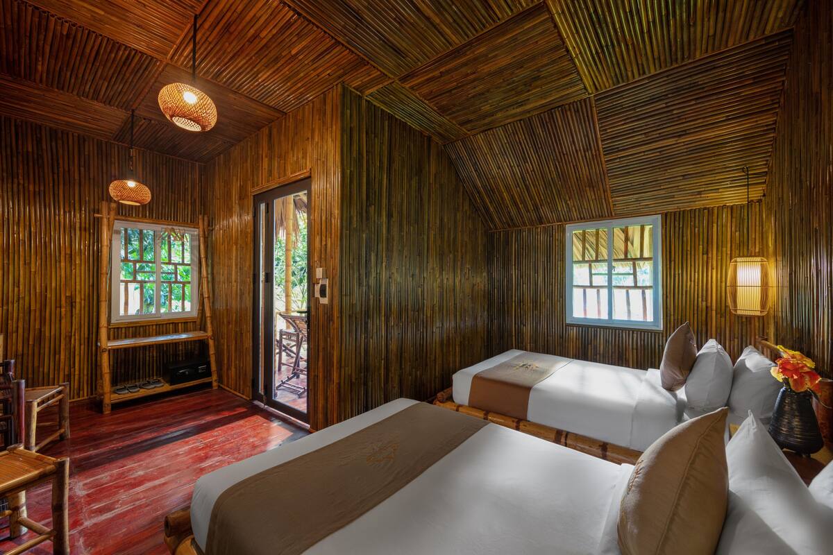Habitación con paredes y suelos de madera, dos camas, una mesita y una ventana con vistas a la vegetación.