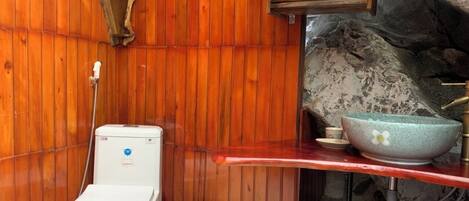 A small bathroom with a wooden wall, a white toilet, a sink with a blue bowl, and a shelf with towels.
