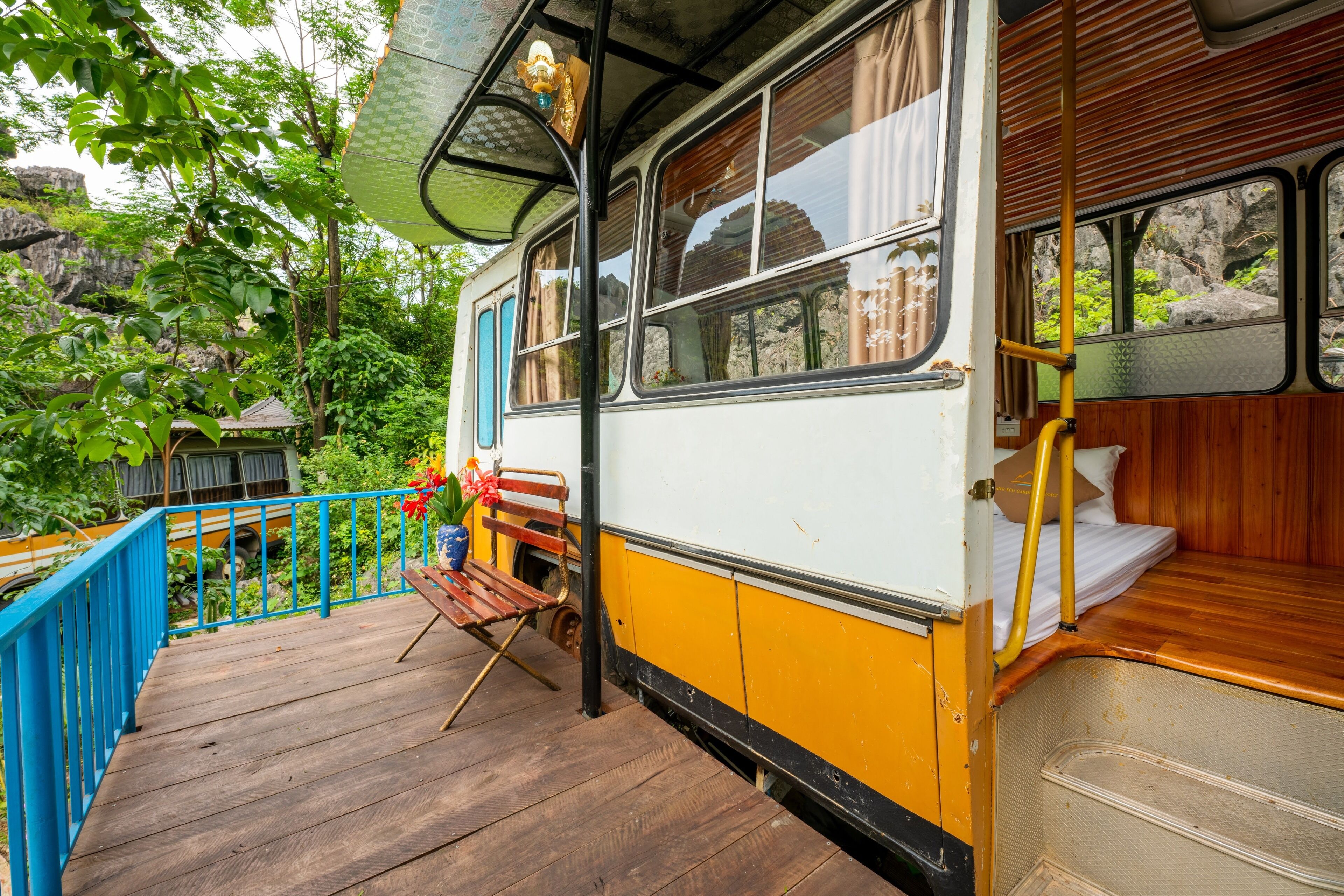 A vintage bus converted into a stationary home with a wooden deck, a bench, and a small garden area.