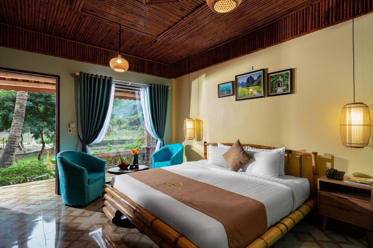 Habitación de hotel con una cama grande, un rincón de descanso con una silla, cabecera de madera, mesita de noche y vistas al exterior.