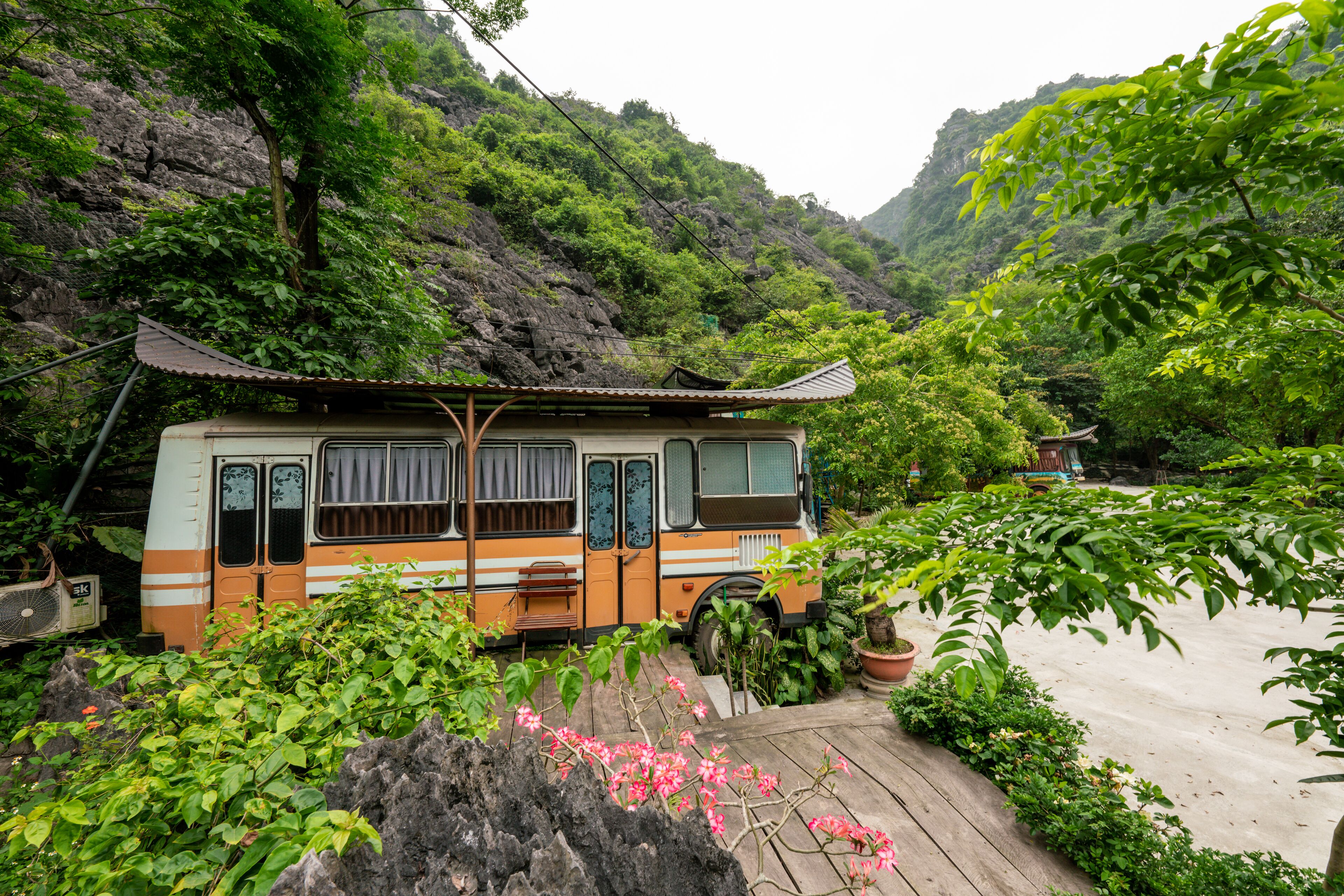 A bus converted into a stationary dwelling with a traditional roof, surrounded by lush greenery and a rocky hillside.