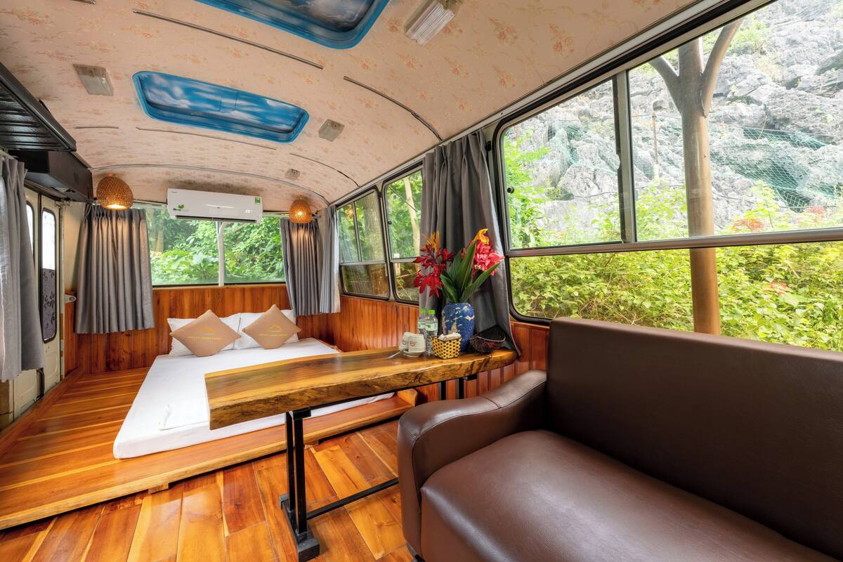Un interior acogedor con suelo de madera, una cama con almohadas, un banco y una ventana con vistas a la vegetación.