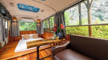 Um interior acolhedor com piso de madeira, uma cama com almofadas, um banco e uma janela com vista para o verde.