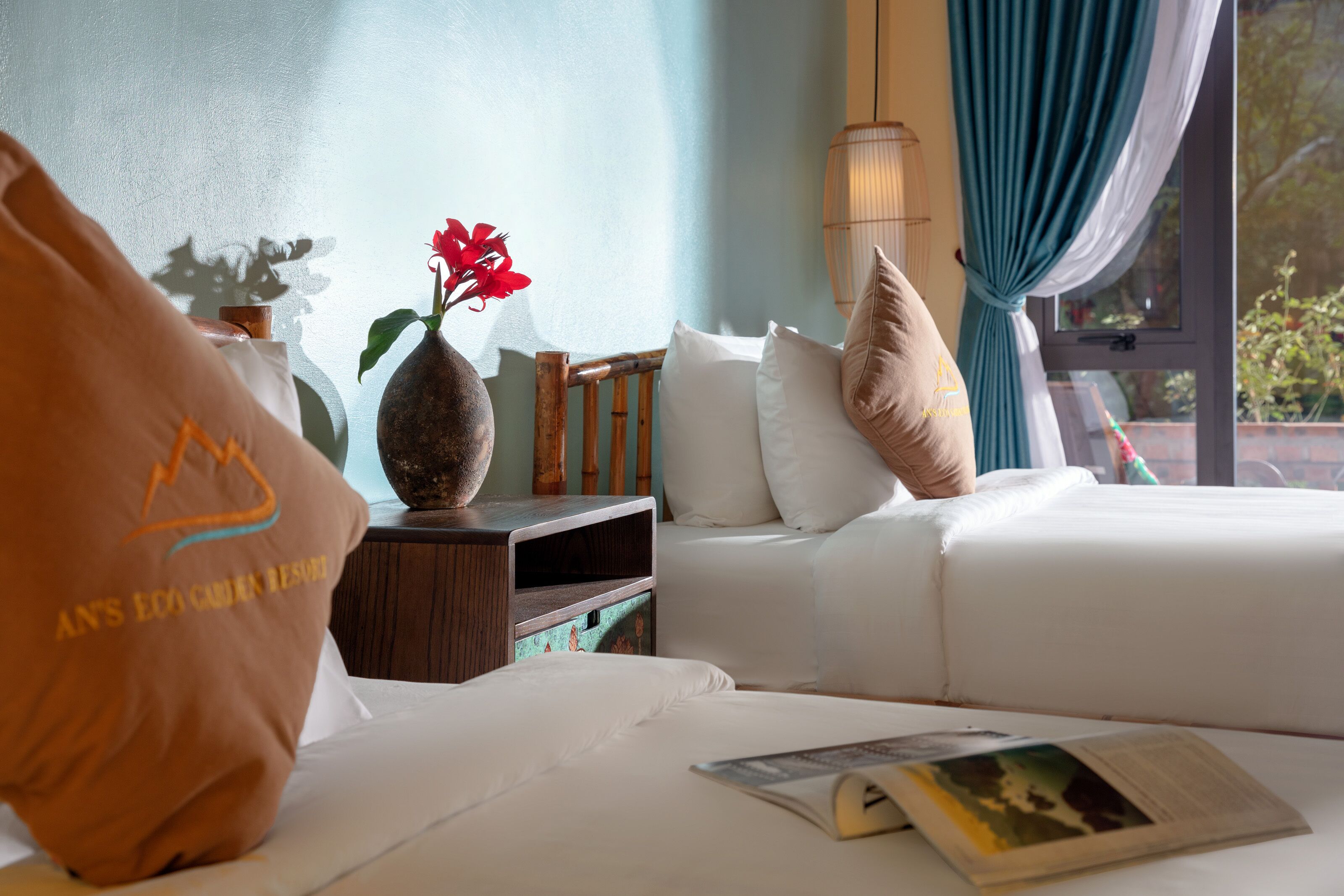 A hotel room with a bed, pillows, a vase with flowers, a bedside table, and a window with curtains.