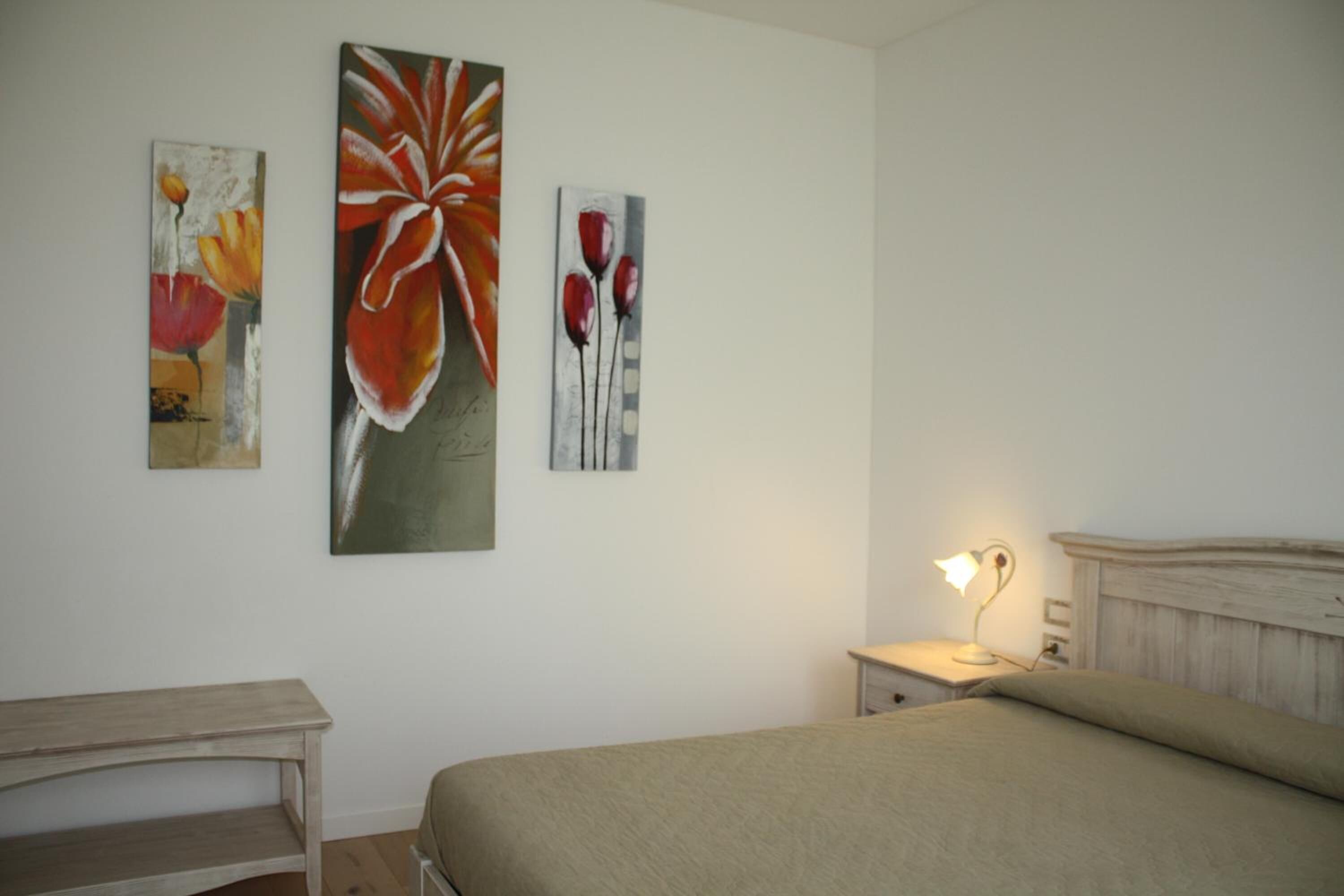 A bedroom with a bed, bedside table, lamp, and three paintings on the wall.