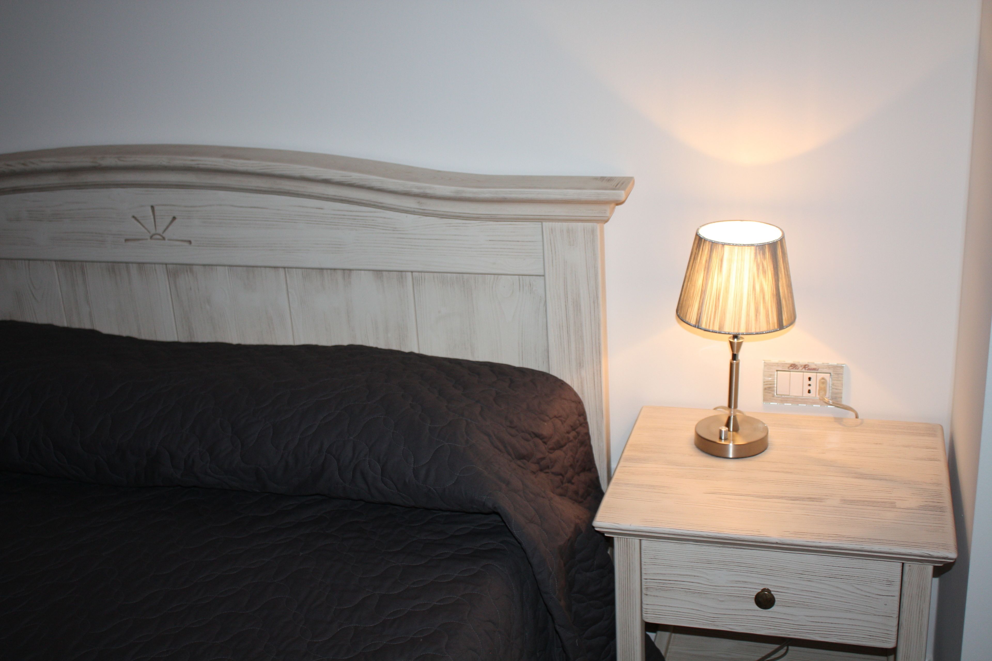 A bed with a dark quilt, a bedside table with a lamp, and a wall socket.