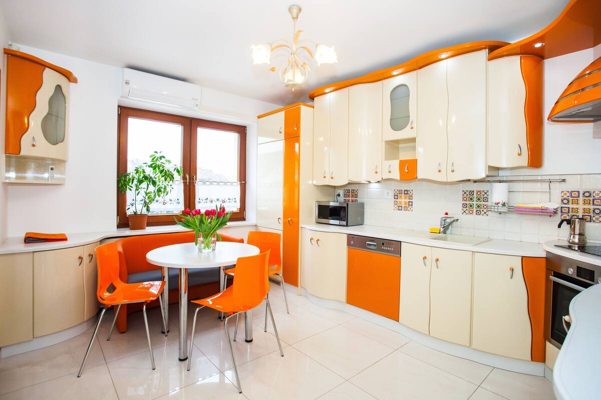 A kitchen with orange and white cabinets, a dining table with orange chairs, and a window with a plant.