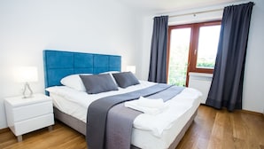 A modern bedroom with a blue headboard, a white bedside table, and a large window with curtains. - Apartamenty SaltApart (Wieliczka)