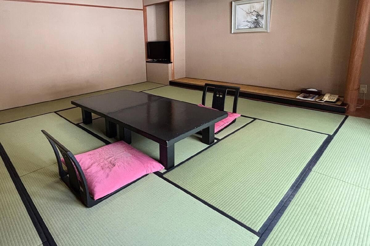 Una habitación con tatami, una mesa baja y sillas, un estante con objetos y un cuadro en la pared.
