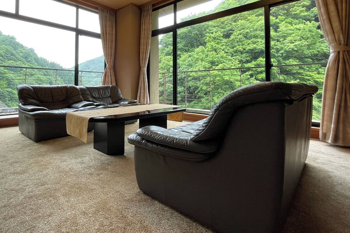 Una habitación con sofá, sillón y mesa de centro, ventanas amplias con vistas a un área boscosa.