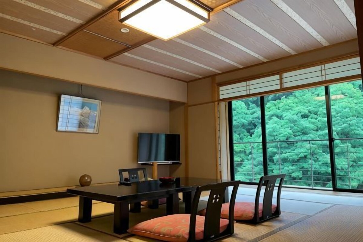 Una habitación japonesa tradicional con suelo de tatami, una mesa baja con cojines, un televisor y un ventanal con vistas a zonas verdes.
