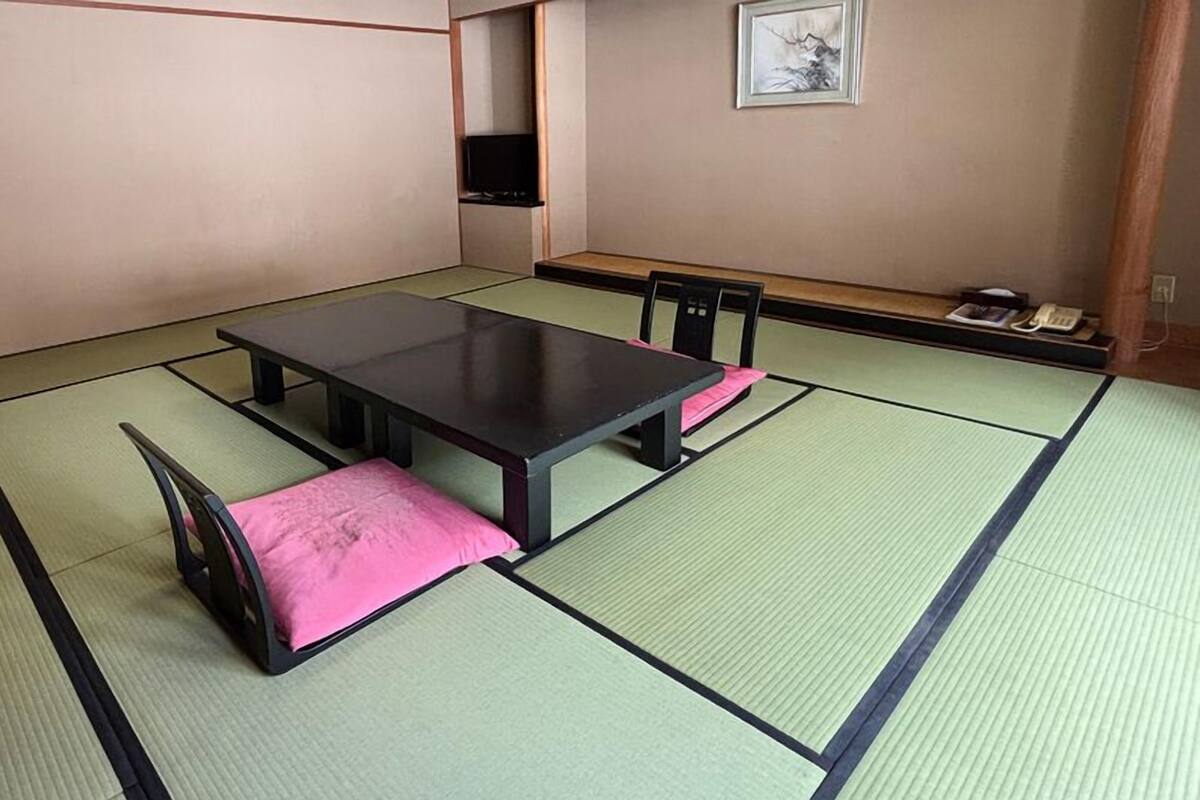 Una habitación con tatami, una mesa baja y sillas, un estante con objetos y un cuadro en la pared.