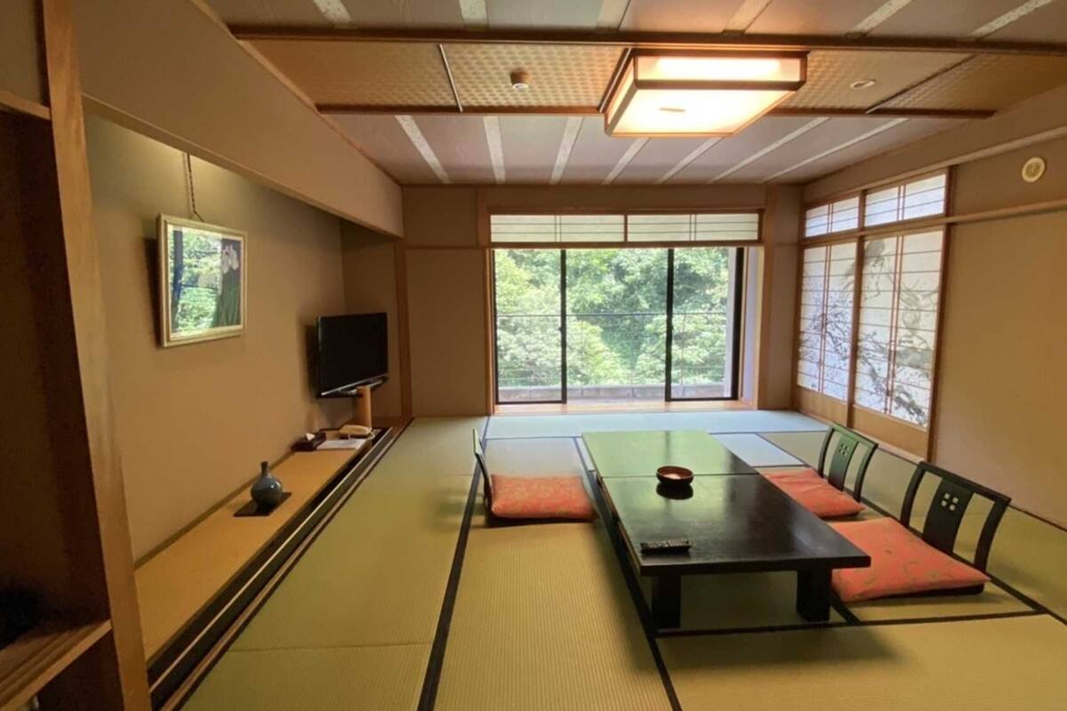 Una habitación con esteras de tatami, una mesa baja, cojines, un televisor y ventanas grandes.