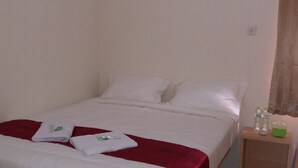 Una cama con sábanas blancas, una manta roja y una mesita de noche de madera con artículos de aseo.
