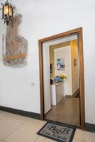 A hotel reception area with a wooden frame door, a wall-mounted light fixture, a bulletin board with various notices, and a doormat with the hotel's name.