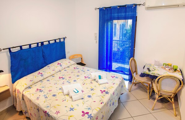 A bedroom with a bed, a chair, a table with items, and a window with blue curtains.