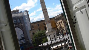 Vista a través de un vano de puerta que muestra una plaza pública con un monumento alto, un edificio con un mural y un cielo despejado.
