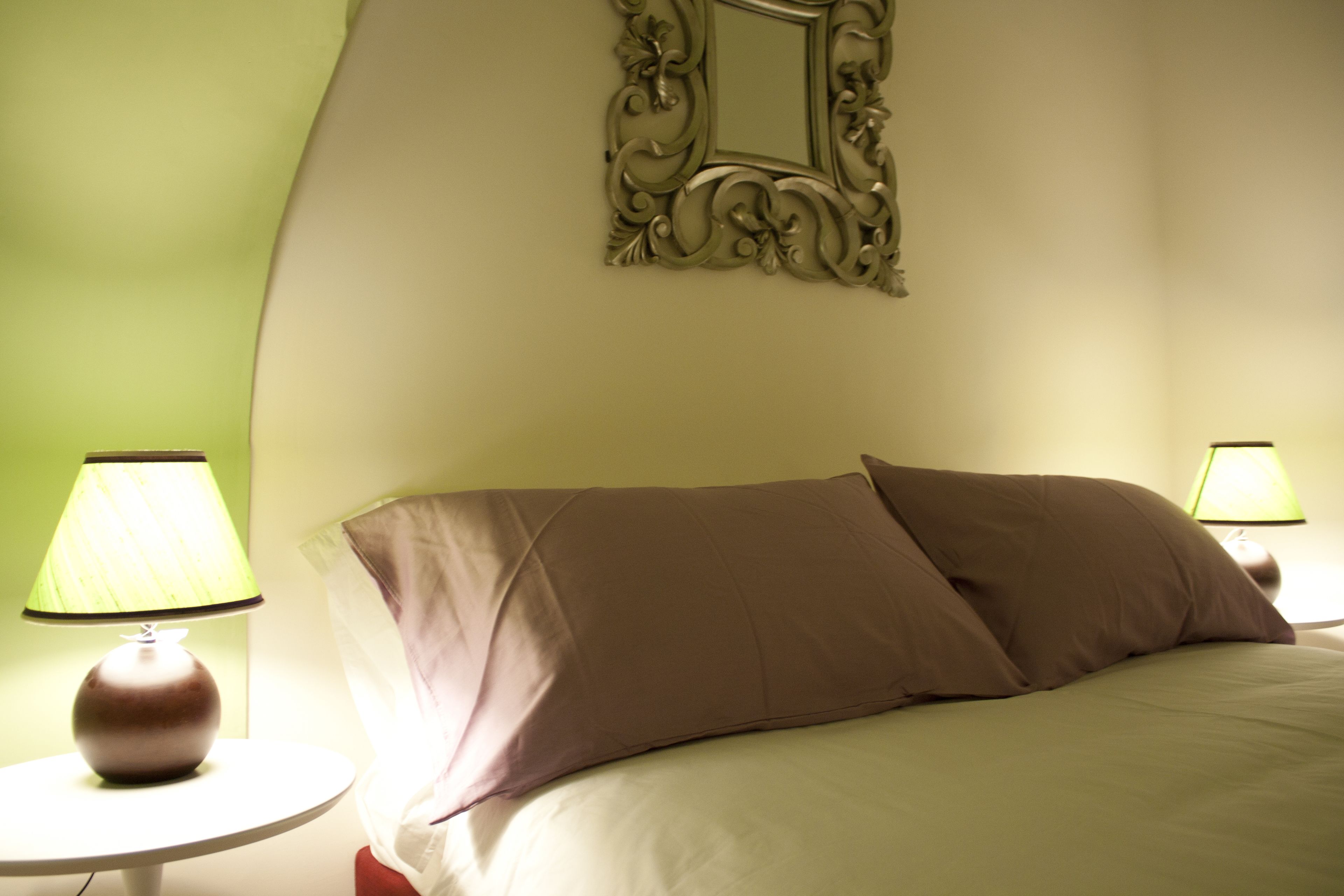 A neatly made bed with two pillows, a round bedside table with a lamp, and a mirror on the wall.