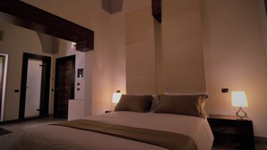 Habitación de hotel con una cama grande, dos mesitas de noche con lámparas, una viga de techo de madera y una puerta que da a otra habitación.
