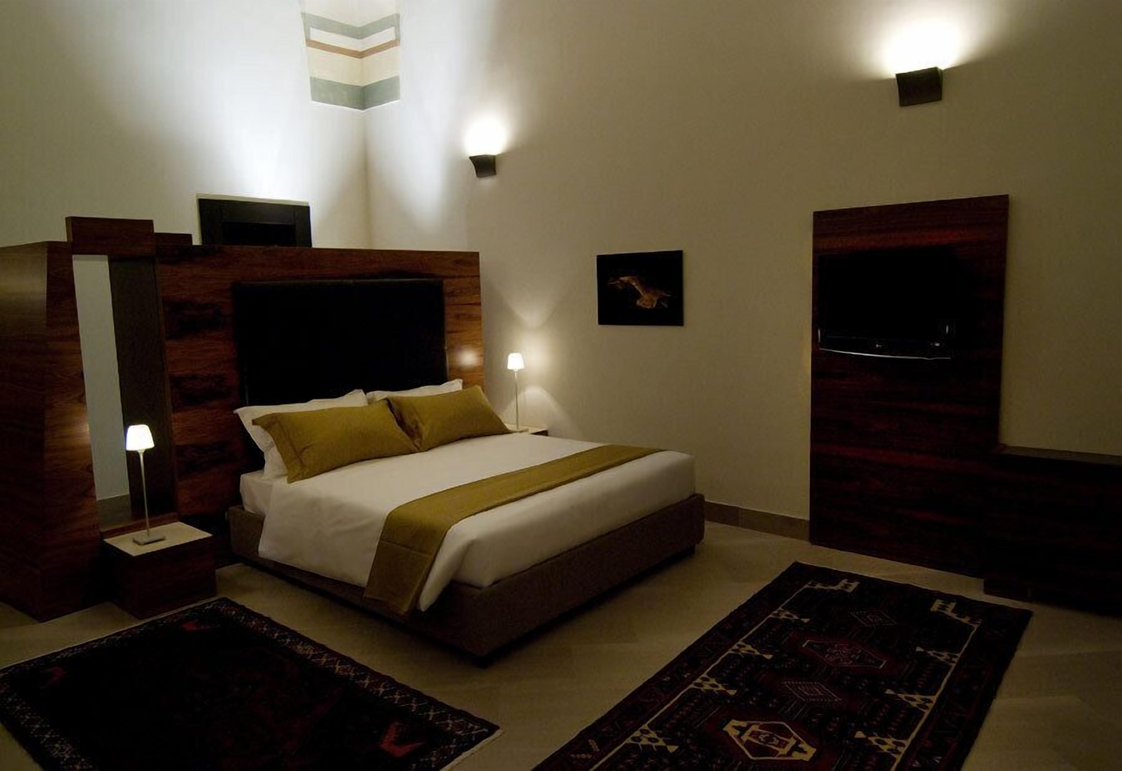 Una habitación de hotel moderna con una cama grande, un televisor de pantalla plana, un cabecero de madera, una mesita de noche con lámpara y una alfombra estampada.