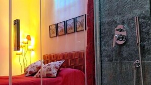 Un dormitorio moderno con un separador de vidrio, una cama roja, ducha y una lámpara fijada a la pared.