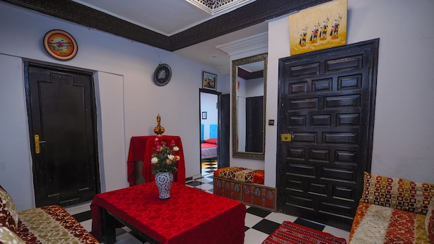 Una habitación con piso a cuadros, una mesa roja con un florero, un sofá con cojines estampados y una puerta negra con un espejo decorativo.