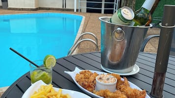 Une table à manger avec une assiette de frites, un bol de viande et de légumes, et une petite coupelle de sauce, le tout placé devant une piscine, avec des bâtiments en arrière-plan.