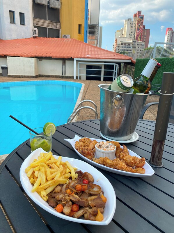 Una mesa de comedor con una fuente de papas fritas, un tazón de carne y verduras, y un recipiente pequeño de salsa, con vista a una piscina y edificios al fondo.