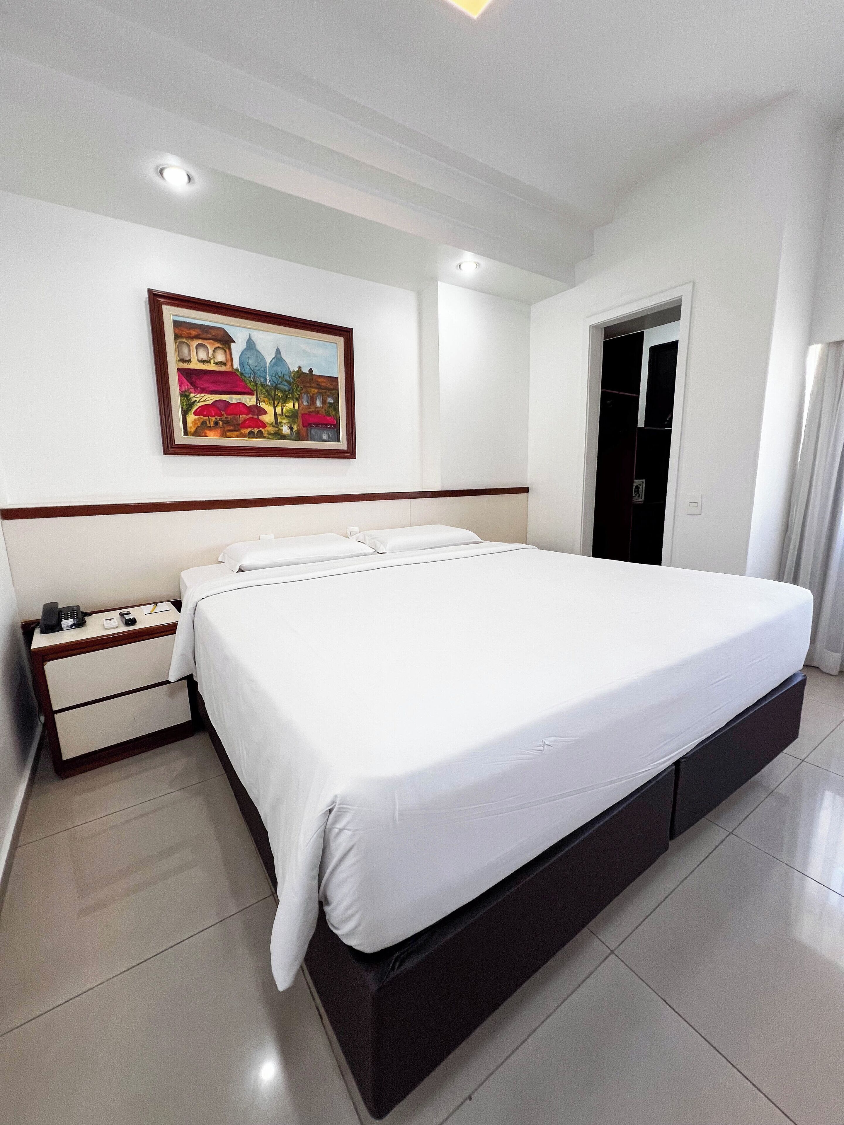 A hotel room with a large bed, a bedside table with a phone, and a painting on the wall.