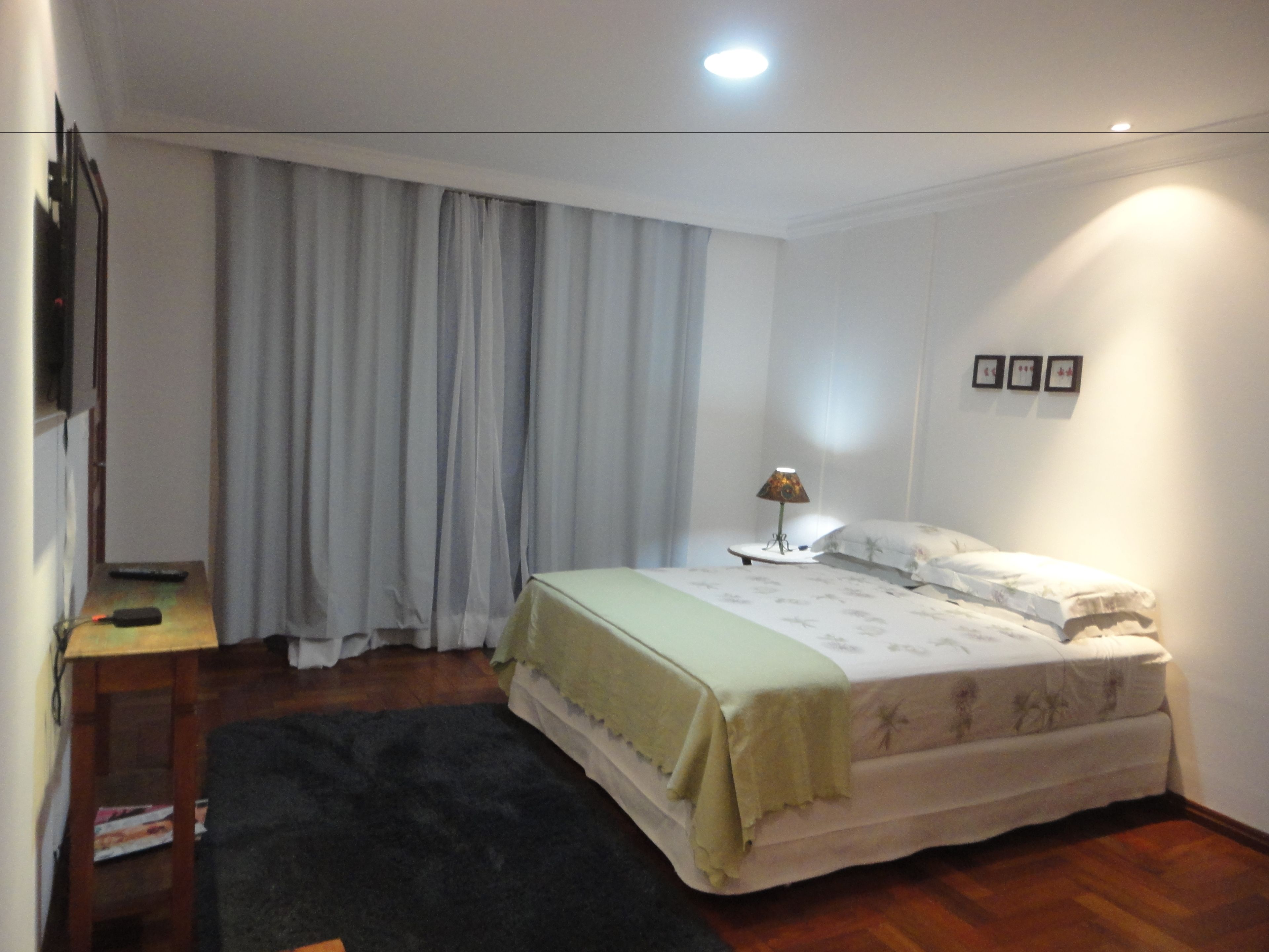 A bedroom with a bed, a nightstand, a lamp, a window with curtains, and a wall-mounted picture.
