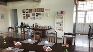 Un comedor con una mesa de madera dispuesta para cuatro personas, rodeada de sillas. Hay cuadros en la pared, una zona de cocina al fondo y una ventana con persianas.