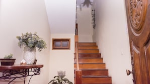 Una escalera de madera con una pared blanca y un ventanal pequeño. A la izquierda, hay una planta en una maceta sobre una mesa.