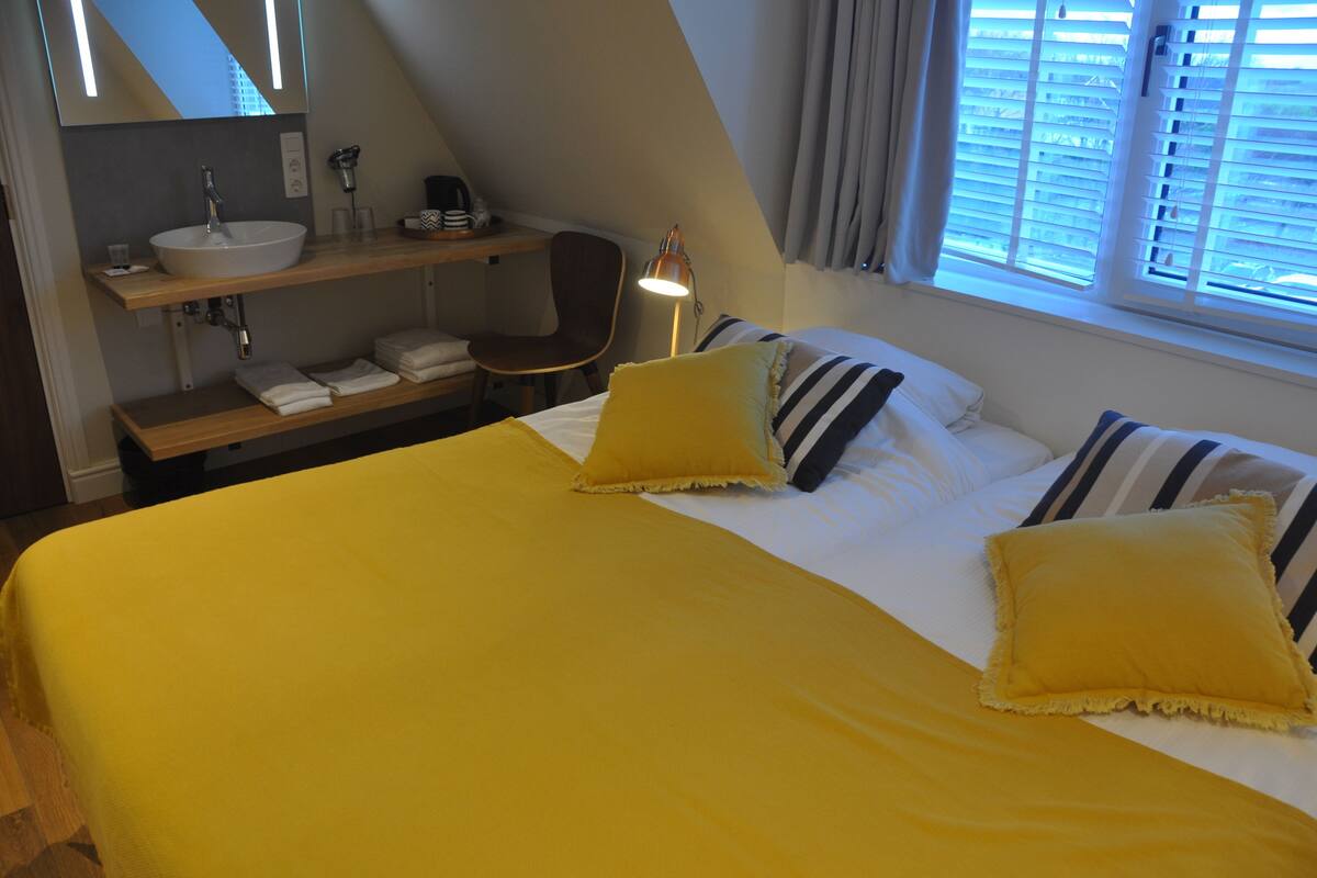 Een slaapkamer met een bed, kussens, een nachtkastje, een lamp, een stoel, een plank met handdoeken en een raam met zonwering.