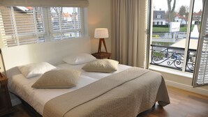 Une chambre à coucher avec un grand lit, une table de chevet avec une lampe, et une vue sur une rue et une rivière, visible par la fenêtre.