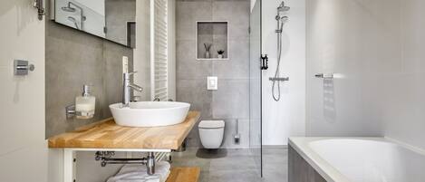 Un baño moderno con una bañera blanca grande, un tocador de madera con lavabo integrado, una ducha con mampara de cristal y un espejo pequeño sobre el lavabo.