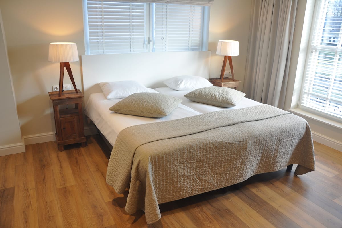 Een net opgemaakt bed met een beige sprei, twee nachtkastjes met lampen, een raam met zonwering en een houten vloer.