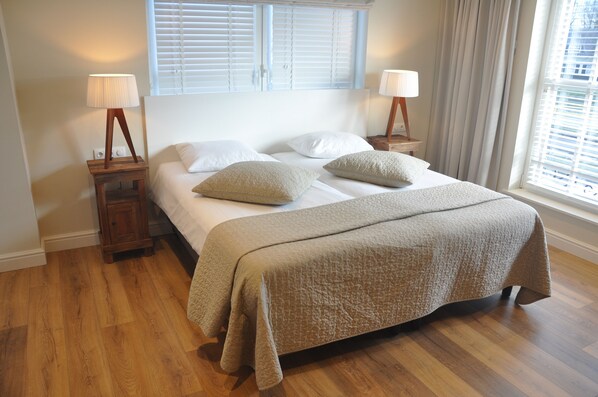 A neatly made bed with a beige quilt, two bedside tables with lamps, a window with blinds, and a wooden floor. - Hotel Sanders de Paauw (Sluis)