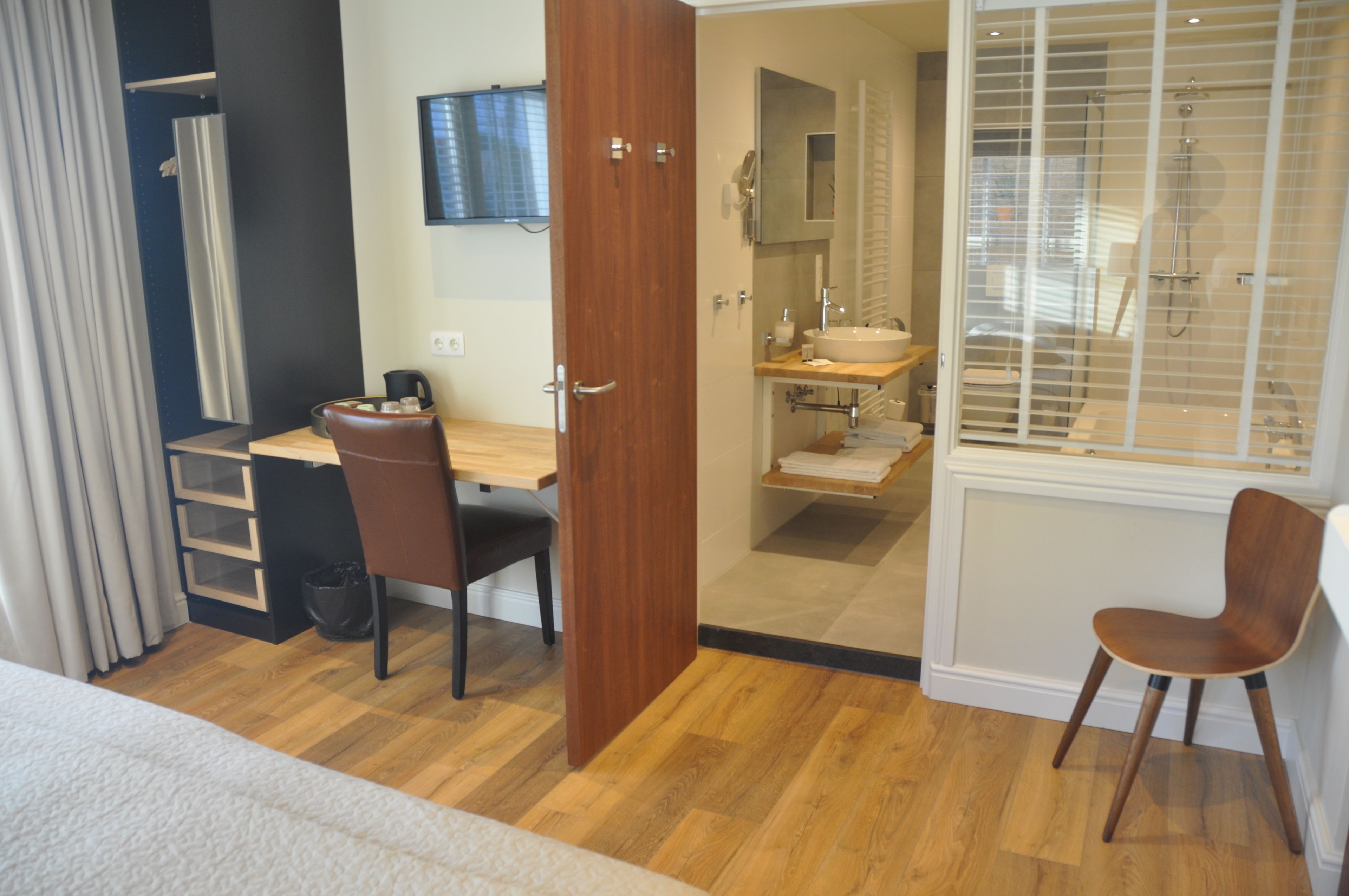 Een hotelkamer met een houten bureau, een stoel, een bed, een badkamer met een wastafel en spiegel, en een douche.