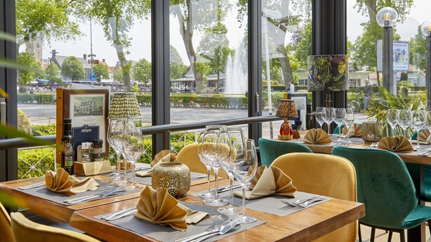 Un restaurant où les tables sont dressées pour le service, offrant une vue sur la rue et les arbres qui se trouvent à l’extérieur.