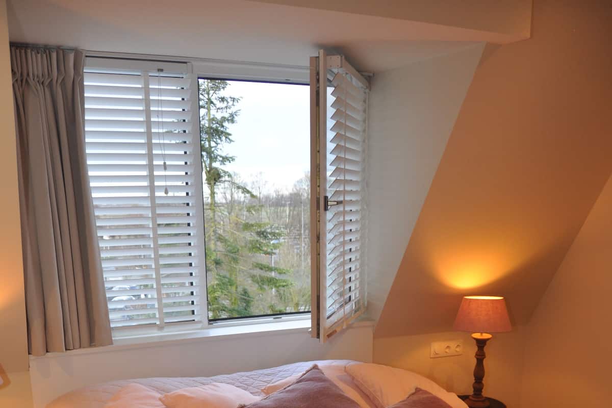 Een slaapkamer met een groot bed, een raam met zonwering, een bedlampje en uitzicht op groen buiten.