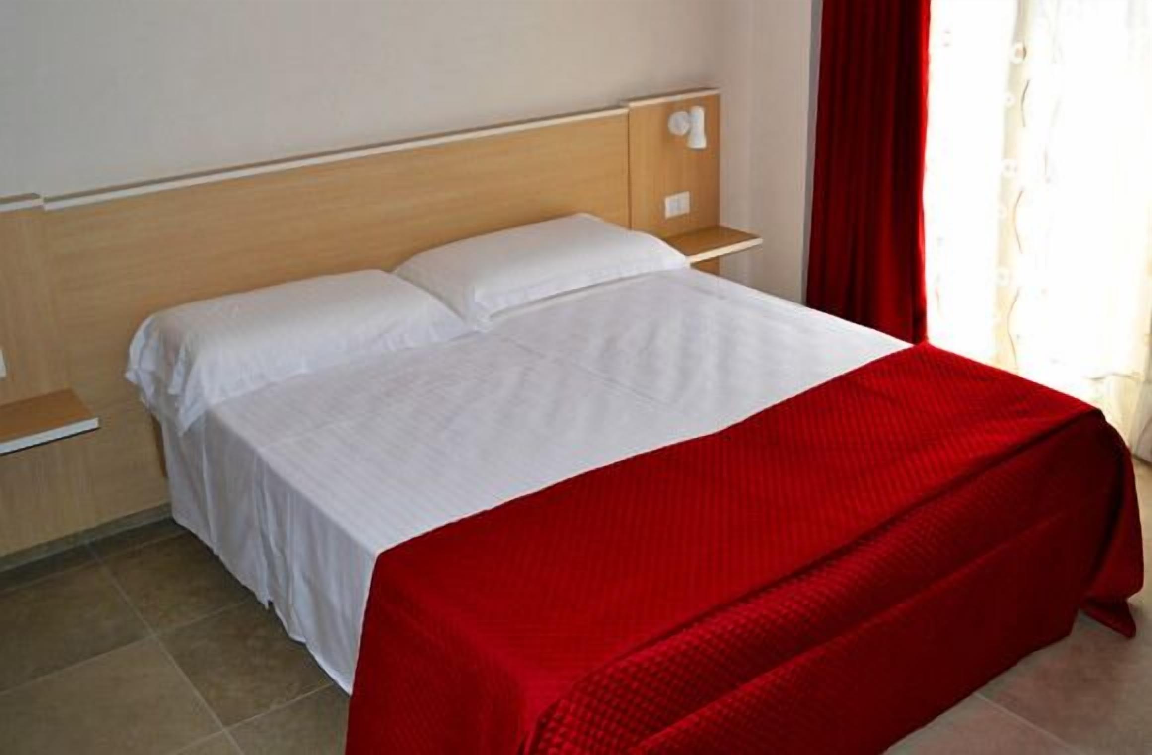 A bed with white linens and a red blanket, a wooden headboard, a nightstand with a white lamp, and a window with red curtains.