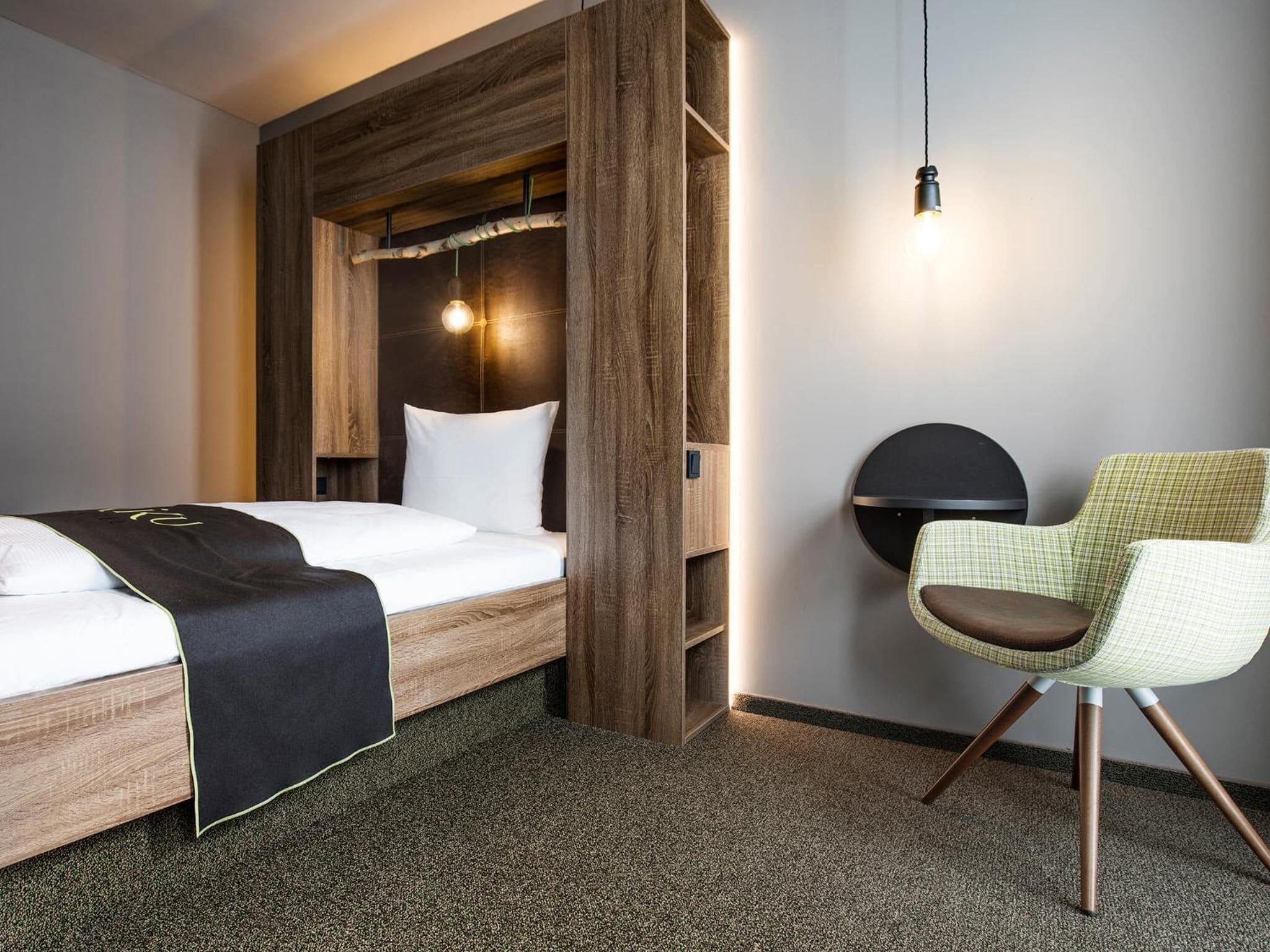 Een moderne hotelkamer met een houten hoofdeinde, een enkel bed met een zwart-wit dekbedovertrek, een rond, aan de muur bevestigd lampje en een geruit fauteuil.