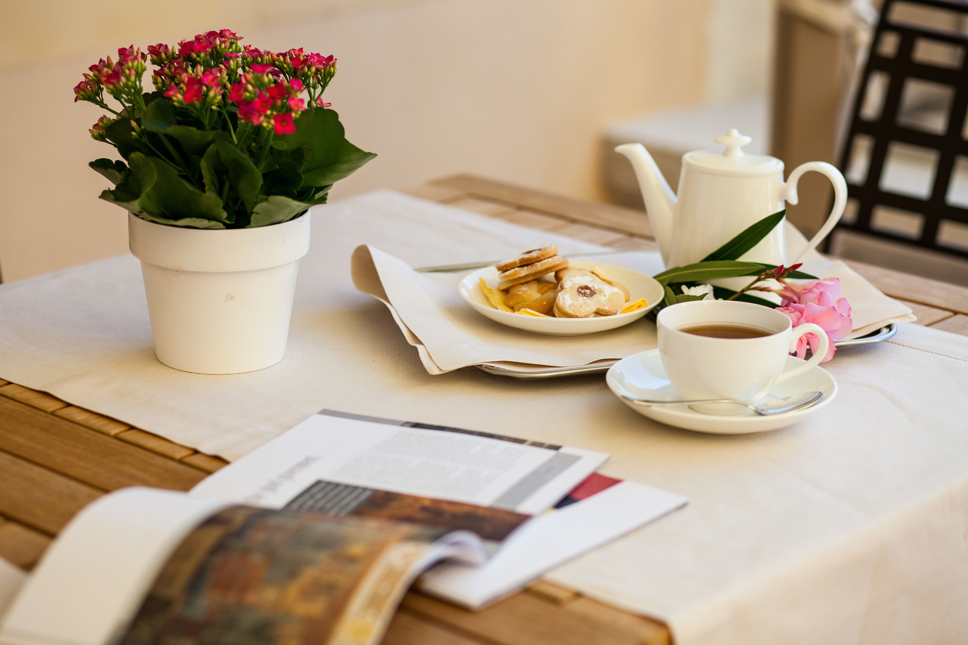 A table with a white teapot, a cup of coffee, a plate with pastries, a small potted plant, and a magazine.