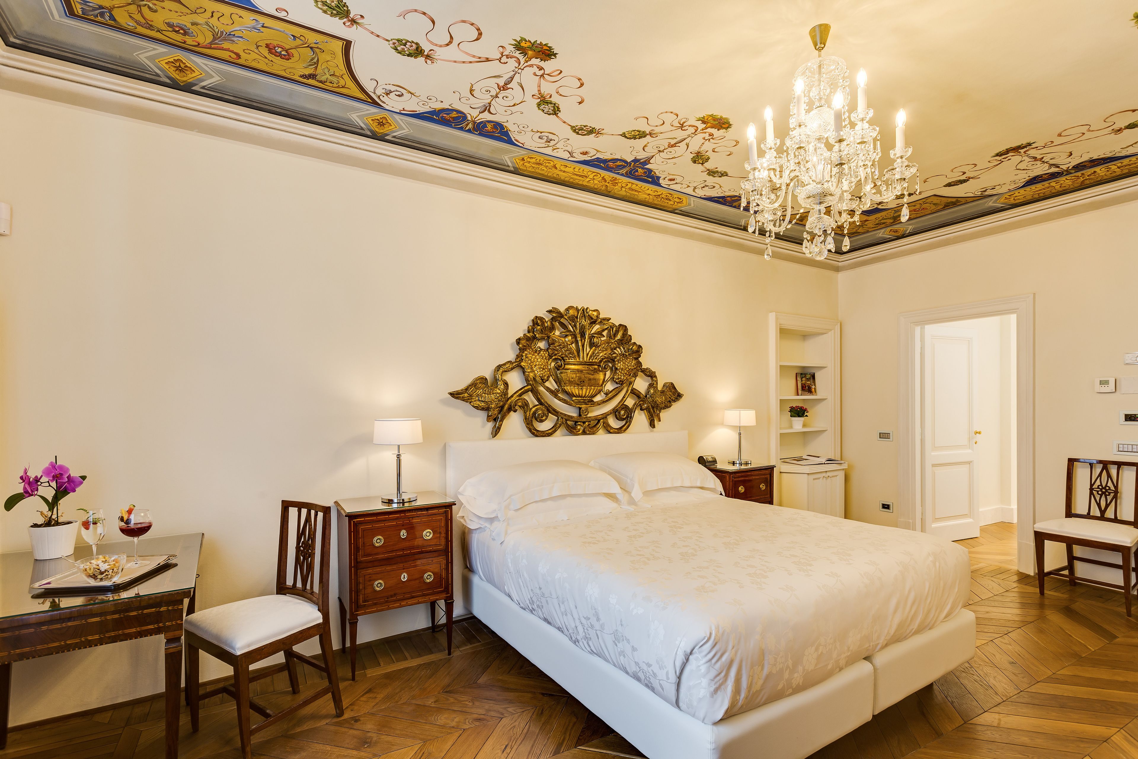 Een slaapkamer met een groot bed, een kroonluchter, een bureau met een stoel en een decoratief plafond.