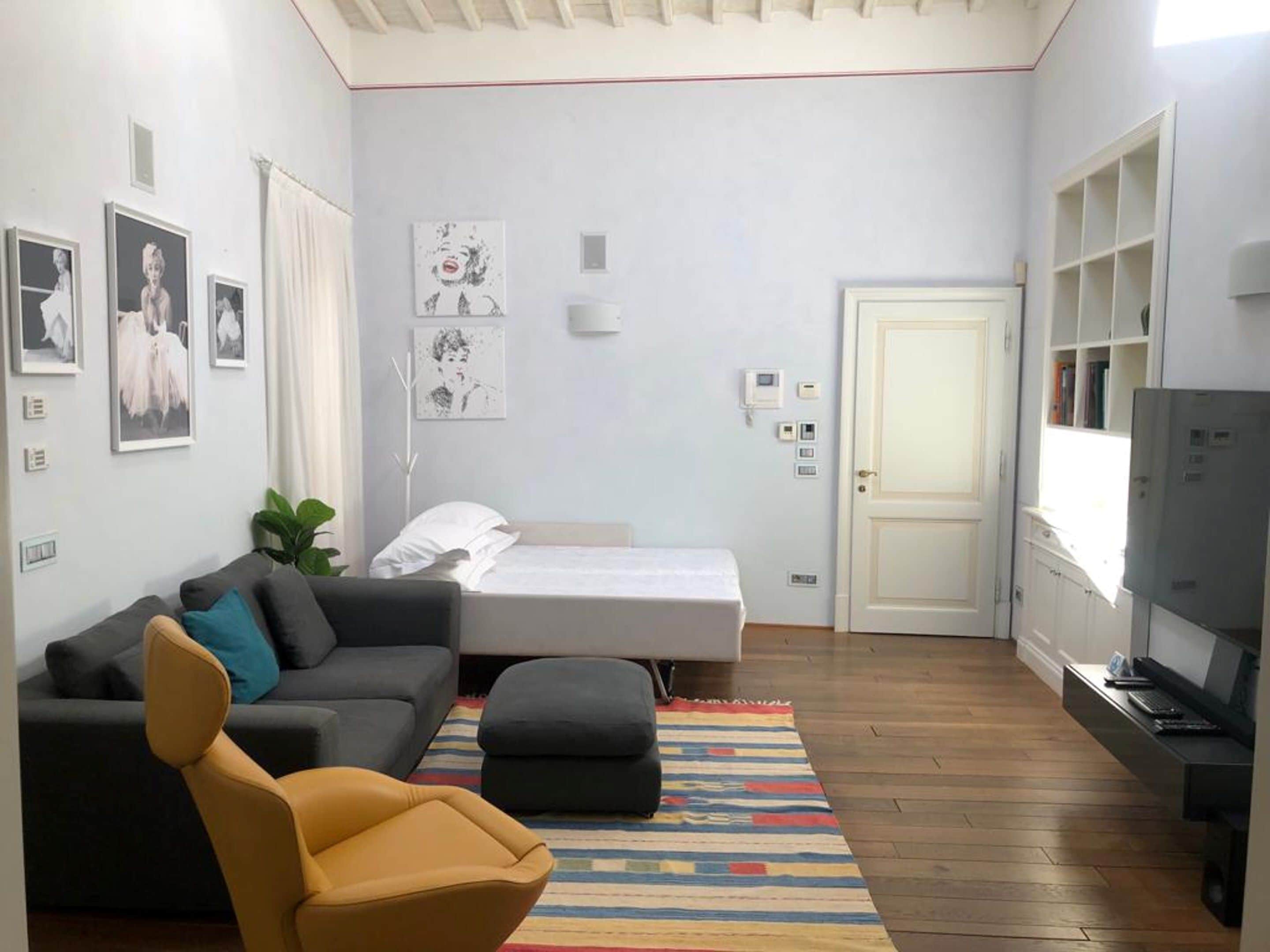 A modern living room with a sofa, ottoman, and a bed. There are framed pictures on the wall, a television, and a bookshelf.