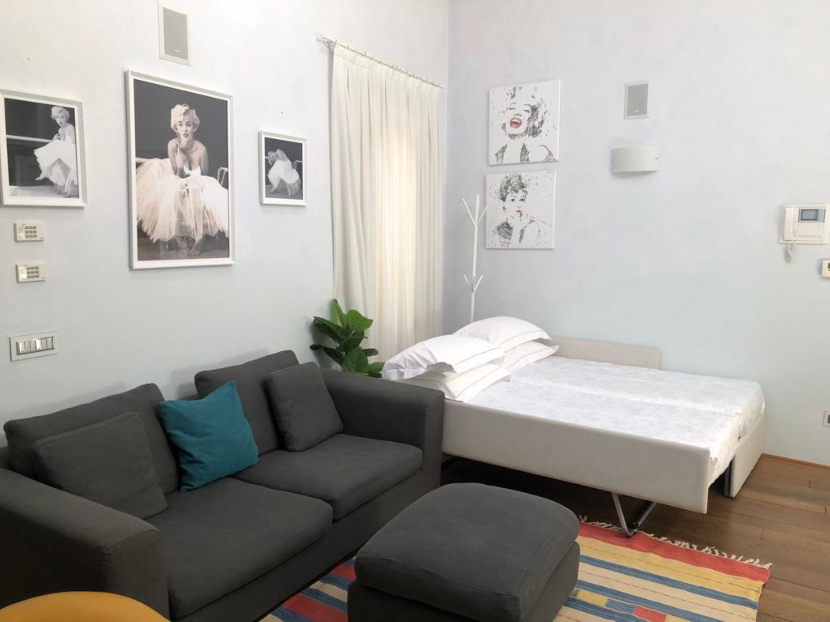 A modern bedroom with a grey sofa, a small ottoman, a bed with white bedding, and a colorful striped rug.