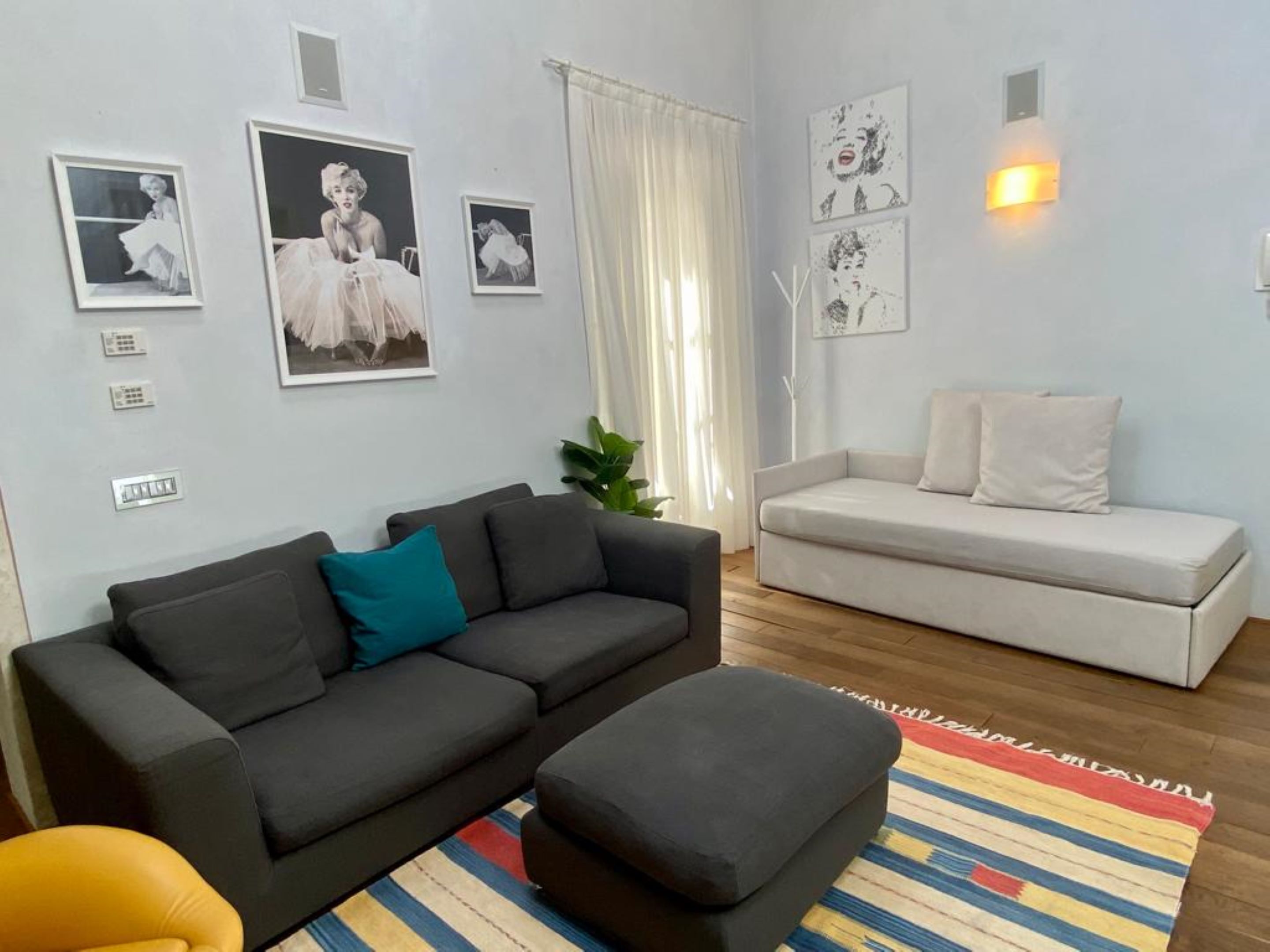 Una sala de estar con un sofá gris oscuro, un sofá blanco y un tapete de rayas de colores. Hay lámparas fijadas a la pared y cuadros enmarcados.