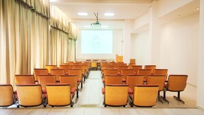 A conference room with rows of orange chairs, a projector screen displaying a hotel logo, and a large window with curtains. - Brivali Hotel e Eventos (Cacador)